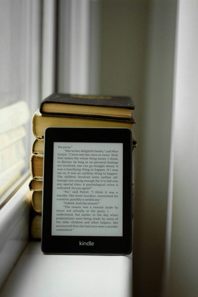 Kindle e-reader sitting on a windowsill next to a stack of books, illustrating e-readers compared for reading